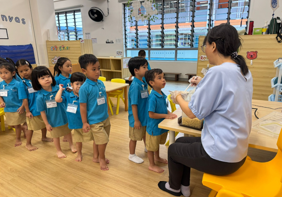 Teacher at table hands food from a tray to a line of children in blue uniforms in a classroom.